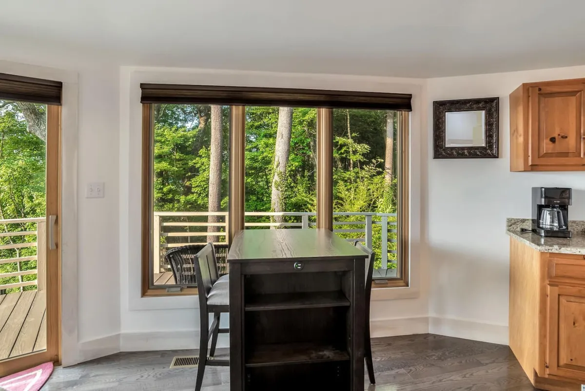 A light-filled dining area with expansive windows overlooking lush greenery, a wooden dining table, and granite countertops. The scene evokes a cozy, intimate atmosphere ideal for morning coffee or a quiet meal while surrounded by nature.