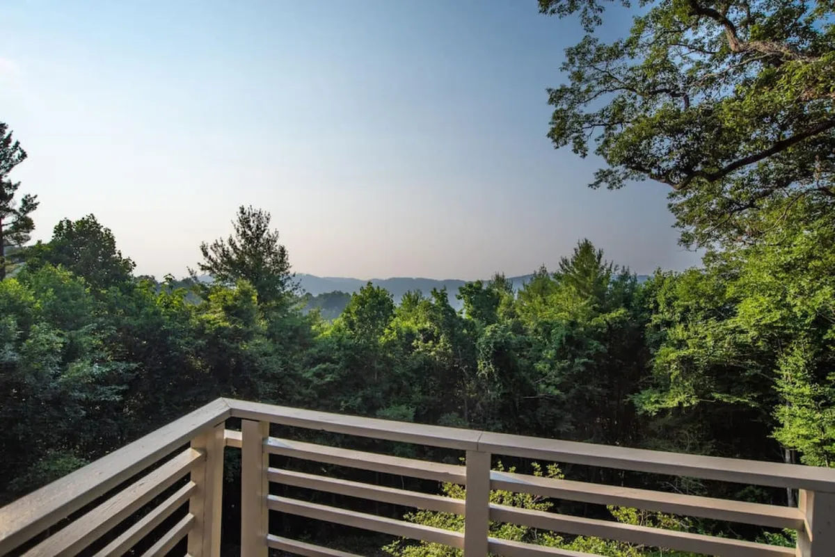 A broad balcony railing overlooks lush green trees and a stunning mountain range in the distance. The scene is quiet and open, inviting contemplation or shared moments on the terrace, with an emphasis on outdoor living and the expanse of the private outdoor area.