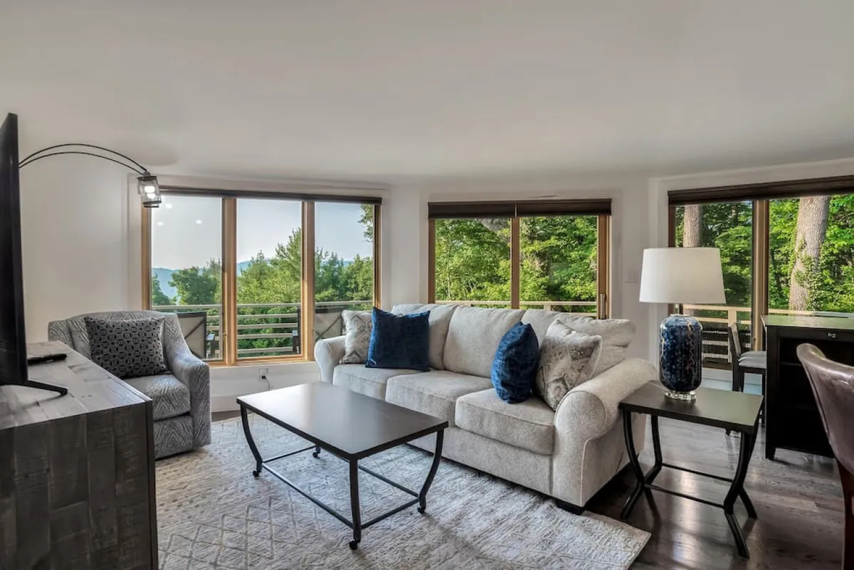 The image showcases a bright, cozy living room with a plush sofa positioned near expansive windows that reveal serene mountain views. A stylish lamp and coffee table create an inviting space for relaxation, while natural light floods the room, highlighting a seamless connection between indoor comfort and the beauty of the outdoors.