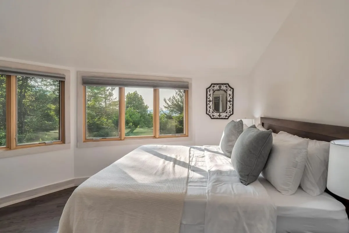 A spacious bedroom featuring a large bed adorned with decorative pillows, flanked by generous windows letting in abundant natural light and offering views of the outdoors. The room’s soothing colors and soft lighting highlight its peaceful, rejuvenating ambiance.