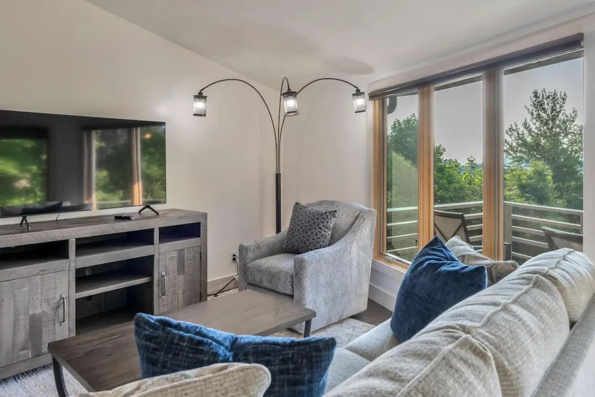A cozy living area featuring a sofa and armchair arranged around a TV. Large windows let in natural light and offer a view of the outdoors, while the layout encourages both relaxation and conversation. The space is inviting, with soft textures and a welcoming ambiance.