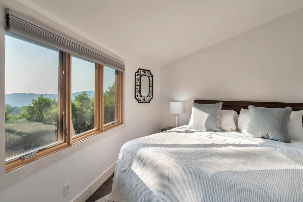 A serene bedroom with large windows framing mountain views, a bed adorned with decorative pillows, and a mirror reflecting natural light. The inviting setting suggests tranquil mornings and private, romantic evenings while being immersed in the surrounding landscape.