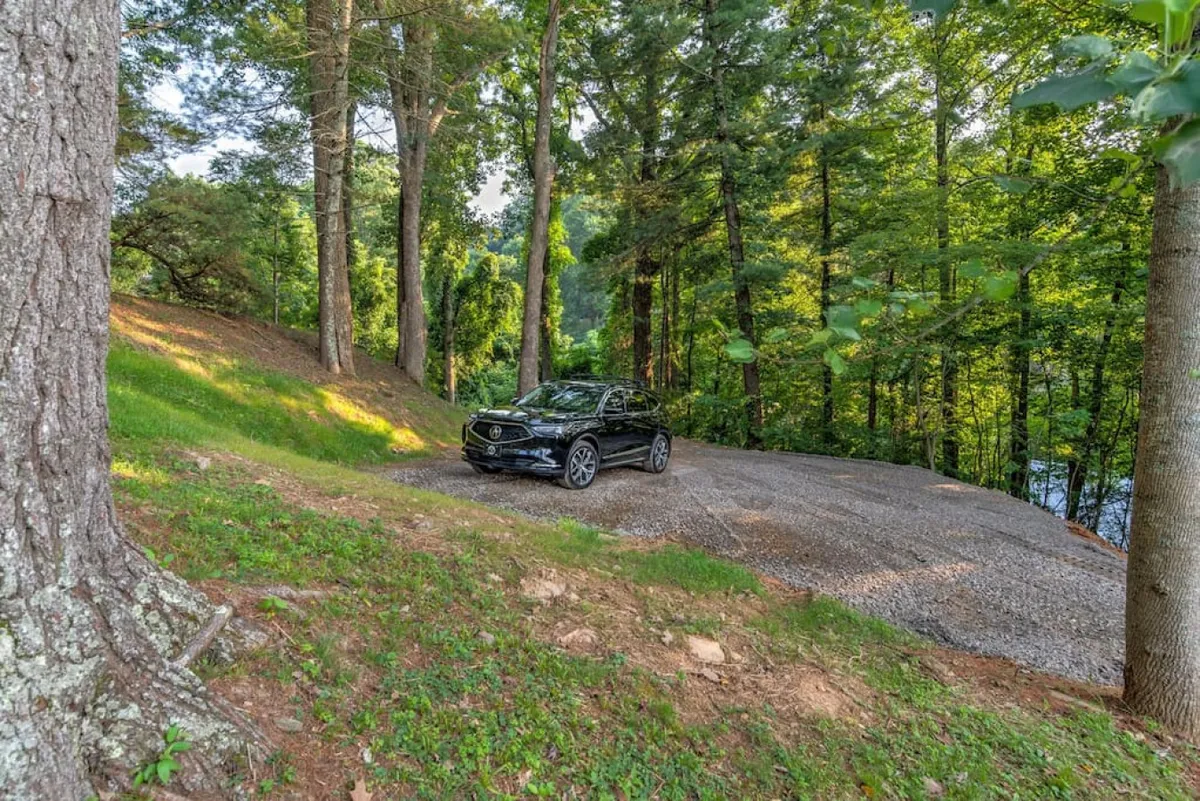An exterior view featuring a black vehicle parked on a gravel driveway surrounded by lush trees, suggesting easy, direct access to the property and a peaceful natural setting.