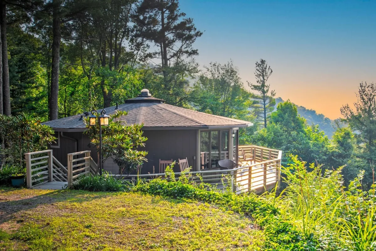 The image displays a unique round structure with extensive decking, surrounded by lush greenery and opening out to a scenic mountain view. The deck is positioned as a social and relaxation space, highlighting possibilities for gatherings, sunrises, and sunsets.