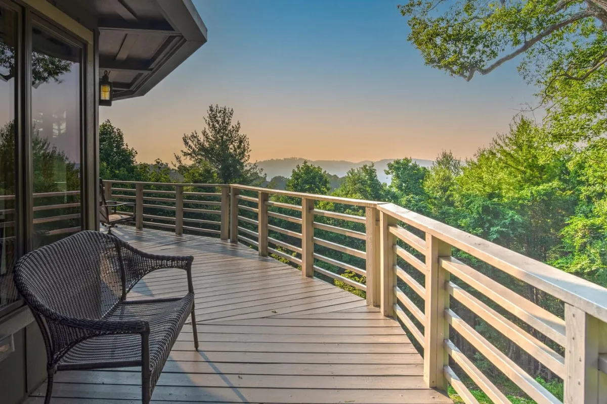 This image features a wooden terrace with a sturdy railing, a comfortable wicker chair, and a dramatic vista of distant mountains. The scene is bathed in natural light, suggesting both morning and evening ambiance. The space evokes a sense of tranquility and highlights the connection between the terrace and the surrounding mountain landscape.