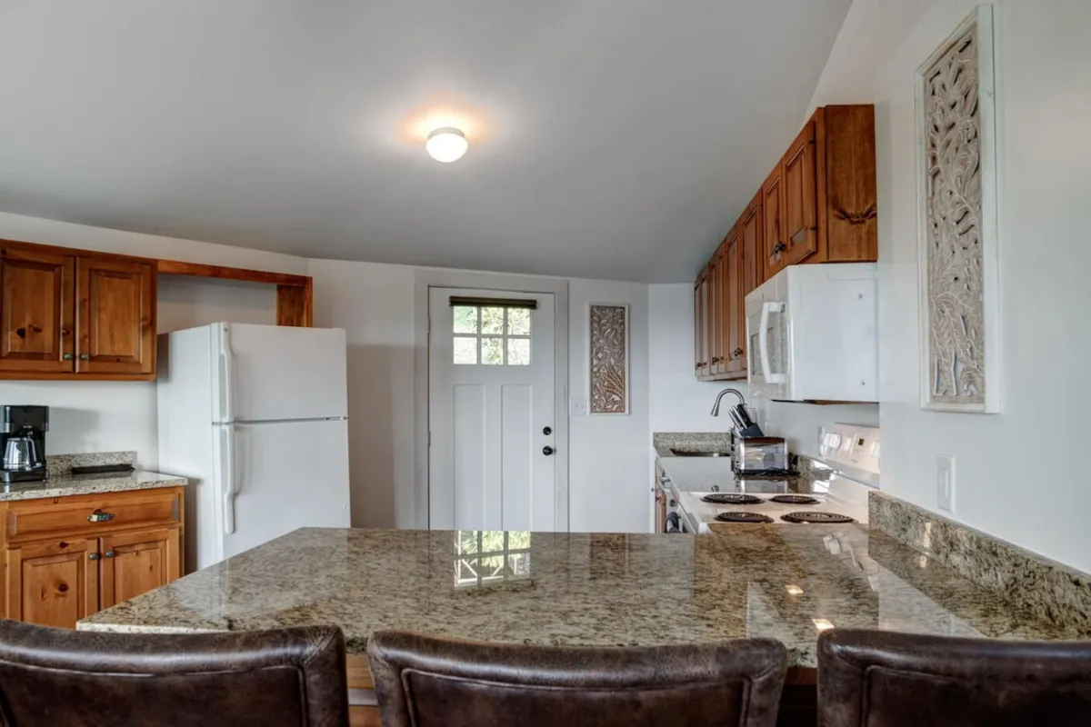 A welcoming interior perspective showing a front door, granite countertop, wooden cabinetry, and white appliances, with enough space to suggest an easy transition from entry into the heart of the cottage.